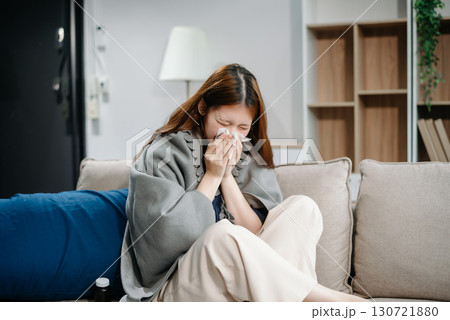 Young Asian woman suffering from flu symptoms, covered with a blanket on a sofa. Concept of illness, healthcare, and recovery. Young Asian woman suffering from flu symptoms, covered with a blanket on a sofa. Concept of illness, healthcare, and recovery. 130721880