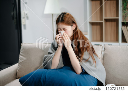 Young Asian woman suffering from flu symptoms, covered with a blanket on a sofa. Concept of illness, healthcare, and recovery. 130721883