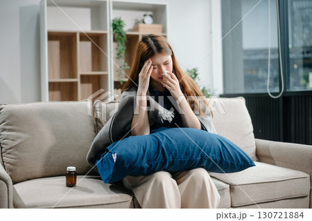 Young Asian woman suffering from flu symptoms, covered with a blanket on a sofa. Concept of illness, healthcare, and recovery. Young Asian woman suffering from flu symptoms, covered with a blanket on a sofa. Concept of illness, healthcare, and recovery. 130721884