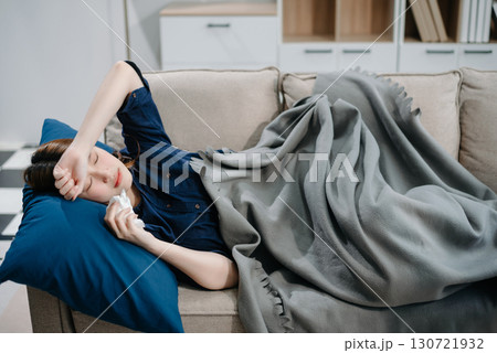 Young Asian woman suffering from flu symptoms, covered with a blanket on a sofa. Concept of illness, healthcare, and recovery. Young Asian woman suffering from flu symptoms, covered with a blanket on a sofa. Concept of illness, healthcare, and recovery. 130721932