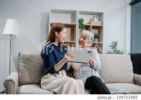 Happy adult Asian granddaughter and senior grandmother having fun enjoying talk sit on sofa in modern living room, using a laptop, tablet 130721995