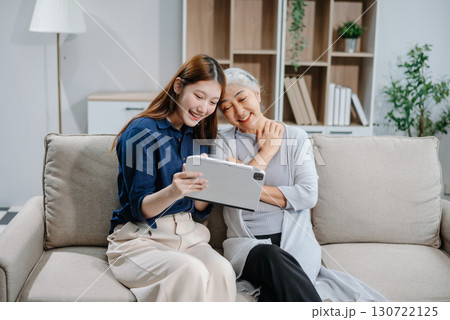 Happy adult Asian granddaughter and senior grandmother having fun enjoying talk sit on sofa in living room using a laptop, tablet 130722125
