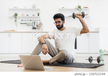 Young adult man sits on exercise mat in home kitchen lifting dumbbells by laptop. Engages in remote fitness routine, demonstrating home workout. Young adult man sits on exercise mat in home kitchen lifting dumbbells by laptop. Engages in remote fitness routine, demonstrating home workout. 130722244
