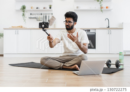 Young adult man recording video blog about fitness and health at home, sitting cross-legged on mat, using smartphone mounted on stick, with laptop and water bottle nearby in bright kitchen setting. Young adult man recording video blog about fitness and health at home, sitting cross-legged on mat, using smartphone mounted on stick, with laptop and water bottle nearby in bright kitchen setting. 130722275