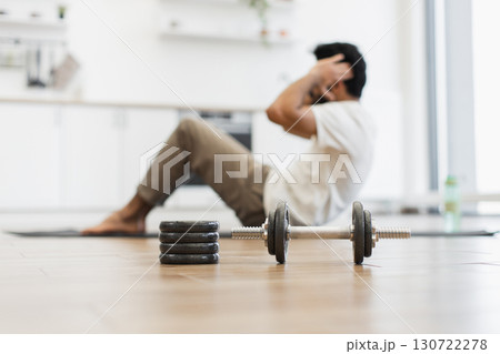 Young adult man indoors doing sit-ups with focus on dumbbell representing fitness theme Young adult man indoors doing sit-ups with focus on dumbbell representing fitness theme 130722278