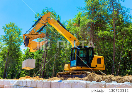 Heavy machinery operates at construction site, lifting concrete block amidst lush greenery under build retaining walls 130723512