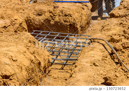 Workers are preparing solid foundation with rebar grid placed in trench filled with earth at construction zone during work day Workers are preparing solid foundation with rebar grid placed in trench filled with earth at construction zone during work day 130723652