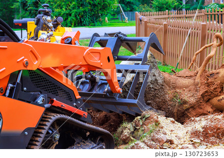 Workers are using compact track loader to extract large tree stump from backyard in suburban neighborhood. Workers are using compact track loader to extract large tree stump from backyard in suburban neighborhood. 130723653