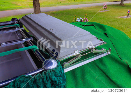 Casket covered with green cloth is situated in calm cemetery, surrounded by blooming flowers on solemn daytime ceremony. Casket covered with green cloth is situated in calm cemetery, surrounded by blooming flowers on solemn daytime ceremony. 130723654