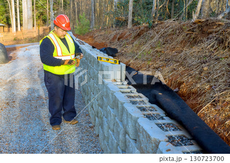 Construction worker wearing safety gear inspects stone retaining wall alignment while working on landscaping project. Construction worker wearing safety gear inspects stone retaining wall alignment while working on landscaping project. 130723700