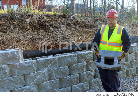 Construction worker in safety vest,helmet lifts block for retaining wall Construction worker in safety vest,helmet lifts block for retaining wall 130723746