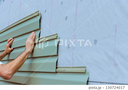 Workers are carefully aligning securing green PVC vinyl siding panels on exterior of home under renovation. Workers are carefully aligning securing green PVC vinyl siding panels on exterior of home under renovation. 130724017