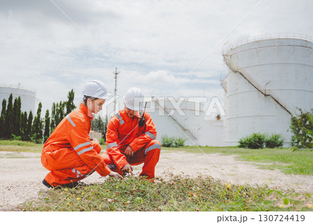Oil and gas engineers inspecting ground vegetation near refinery tank in safety gear and helmets 130724419