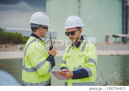 Two engineers using radio and tablet at coastal oil gas facility in safety jackets 130724421