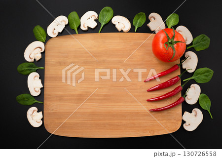 A blank wooden cutting board with a variety of fresh ingredients like mushrooms, spinach, tomatoes, and chili peppers on a black background. A blank wooden cutting board with a variety of fresh ingredients like mushrooms, spinach, tomatoes, and chili peppers on a black background. 130726558