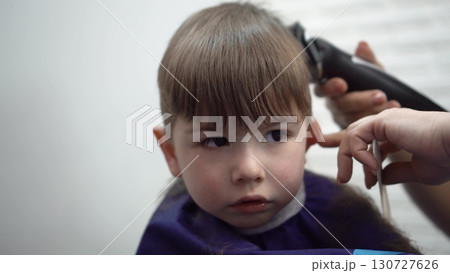 a three-year-old child curiously watches himself in the mirror while a barber gently cuts his hair in a cozy kids salon 130727626