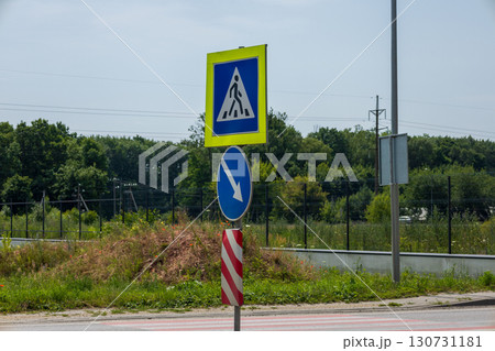 Blue pedestrian crossing sign and a downward arrow sign on a red white striped pole. Roadside with green grass, lush trees, clear blue sky. Blue pedestrian crossing sign and a downward arrow sign on a red white striped pole. Roadside with green grass, lush trees, clear blue sky. 130731181