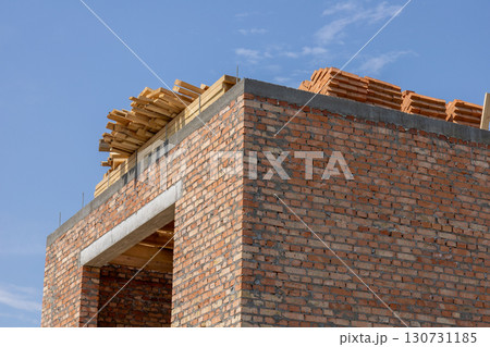 Developing house construction project showing unfinished red brick walls and wooden roof framing. Stacked building materials under a clear blue sky. Developing house construction project showing unfinished red brick walls and wooden roof framing. Stacked building materials under a clear blue sky. 130731185