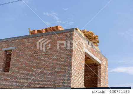 New residential brick house construction site. Unfinished building exterior with wooden roof beams and stacked tiles. Clear blue sky backdrop. 130731186