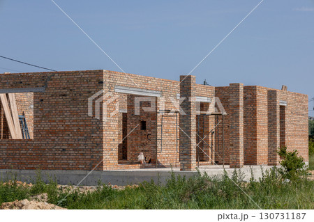 Unfinished new home construction with red brick walls and concrete foundation on a building site under a bright blue sky. 130731187