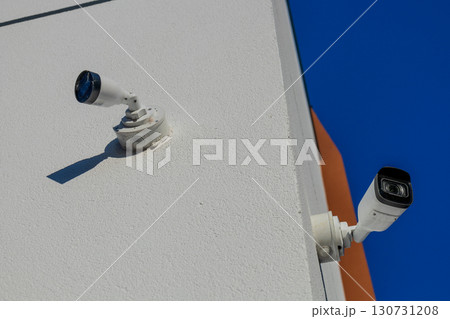 White security camera and outdoor light fixture mounted on a textured building wall. Clear blue sky, bright sun, long shadows. 130731208