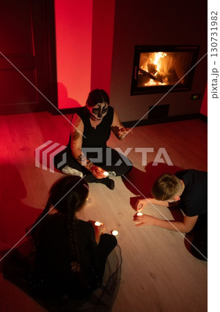A spooky Halloween gathering with a skull-faced adult and two children lighting candles in a dimly lit room. 130731982