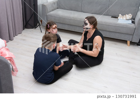 A woman with face paint sits with two children, also with face paint, on the floor. They appear to be engaged in conversation or a game. 130731991