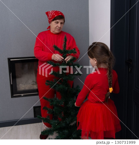 Grandmother and granddaughter decorating a small Christmas tree together, wearing matching red outfits. 130731999