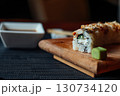 A sushi roll sits on a wooden cutting board with a small green leaf on top 130734120