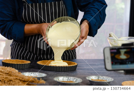 Baker in black and white striped apron pouring creamy custard filling from glass bowl into prepared graham cracker crusts in fluted tart pans kitchen countertop 130734664
