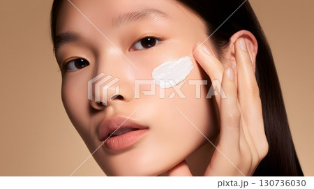 A beautiful face shows radiant skin while a hand gently applies a white beauty cream for healthy and glowing complexion care. 130736030