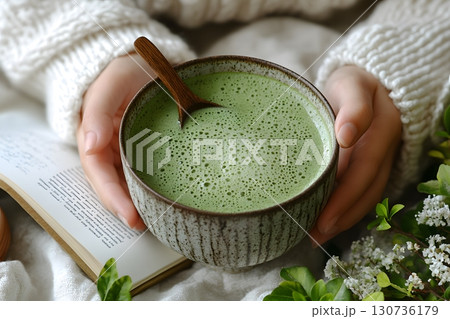 Hands in a soft sweater gently hold a rustic bowl filled with vibrant green matcha, perfect for a calming break. 130736179