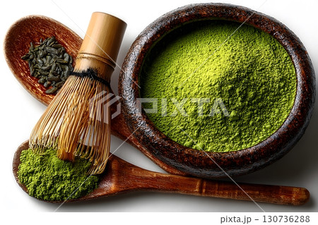 Bright green matcha powder fills a dark wooden bowl, alongside a bamboo whisk and tea leaves on wooden spoons. 130736288