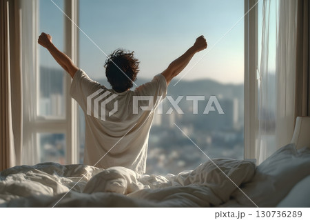 A person rises from bed, stretching arms high towards the sunny window, feeling refreshed, joyful, and optimistic for the promising new day. 130736289