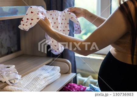 A pregnant woman carefully holds a baby's jumpsuit as she arranges the clothes in a dresser drawer in preparation for the arrival of her newborn. A pregnant woman lovingly sorts through baby clothes. A pregnant woman carefully holds a baby's jumpsuit as she arranges the clothes in a dresser drawer in preparation for the arrival of her newborn. A pregnant woman lovingly sorts through baby clothes. 130736983