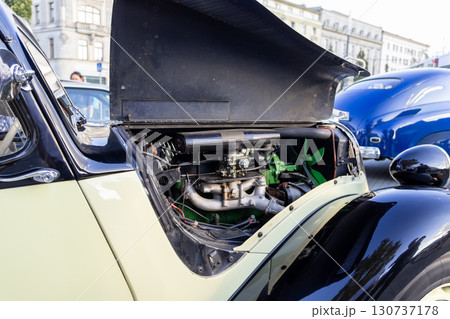 Close view of a vintage beige and black car's open hood, revealing its intricate classic engine with a green fan, displayed at an outdoor show 130737178