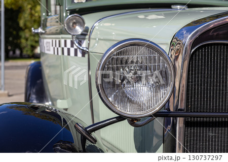 Detailed close-up captures a bright vintage car headlight, its gleaming chrome bezel, distinctive dark grille, and classic checkerboard side panel, embodying automotive history Detailed close-up captures a bright vintage car headlight, its gleaming chrome bezel, distinctive dark grille, and classic checkerboard side panel, embodying automotive history 130737297