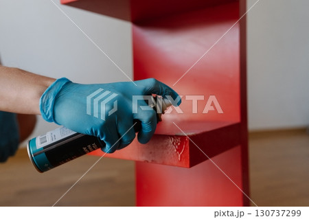 Male hand wearing blue glove applying scotch remover spray to dried adhesive stain on red shelf, performing thorough housekeeping at home, close-up. Concept of easy housekeeping domestic life. Male hand wearing blue glove applying scotch remover spray to dried adhesive stain on red shelf, performing thorough housekeeping at home, close-up. Concept of easy housekeeping domestic life. 130737299