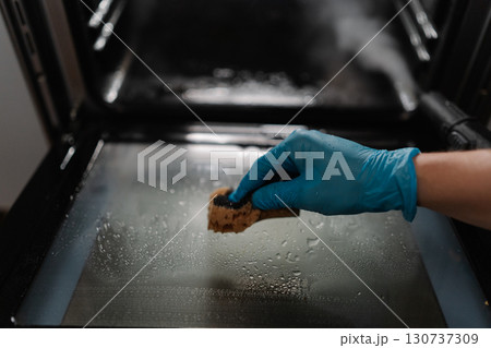 Professional cleaning worker wearing blue glove using sponge for cleaning glass door of gas oven, performing meticulous kitchen appliance maintenance and sanitization, close-up. 130737309
