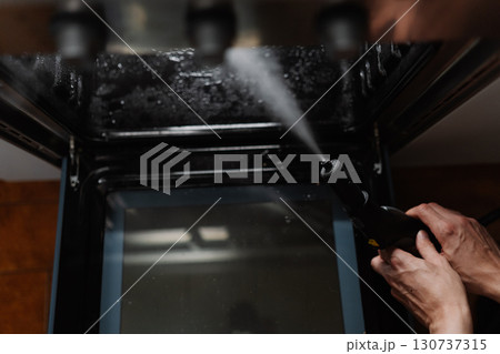 Top view of cleaning person using steam cleaner inside black gas oven, ensuring thorough disinfection and hygiene in kitchen setting, close-up. Concept of hygiene and cleanliness during routine chores 130737315