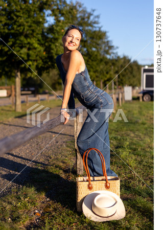 Young adult beautiful woman wear stylish jeans one-shoulder denim jumpsuit have fun walks outdoors warm sunny day holding straw bag hat hand. Female person carefree smiles enjoying countryside nature Young adult beautiful woman wear stylish jeans one-shoulder denim jumpsuit have fun walks outdoors warm sunny day holding straw bag hat hand. Female person carefree smiles enjoying countryside nature 130737648