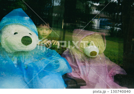 A charming scene featuring two teddy bears dressed in raincoats, and a child peering between them, suggesting a fun and playful moment during a rainy day 130740840
