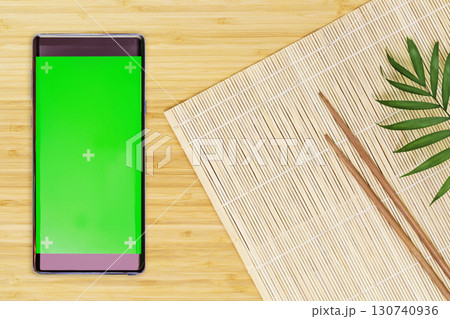 Restaurant Order online concept. Oriental style flat lay with a nature mat, wooden chopsticks and smartphone with green screen on a wooden board. Eco friendly Mock up for online food market, caffee 130740936