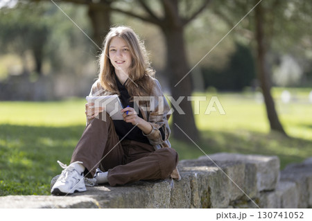 Young woman taking notes in a notebook while sitting on a stone wall in a park 130741052