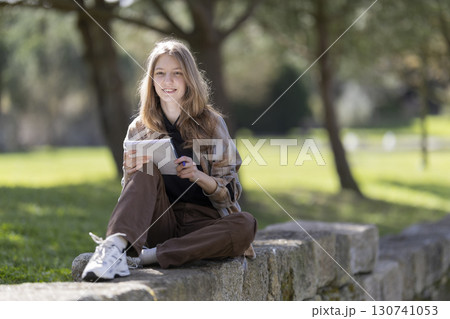 Young woman taking notes in a notebook while sitting on a stone wall in a park 130741053