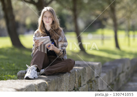 Young woman writing in notebook sitting on stone wall in park 130741055