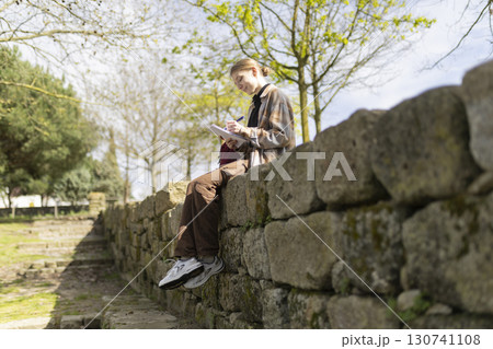 Young woman writing in notebook sitting on stone wall in park Young woman writing in notebook sitting on stone wall in park 130741108