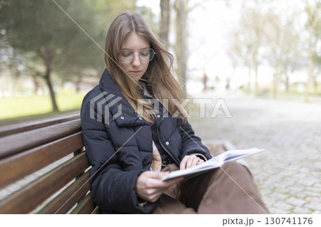 Young woman reading a book on a park bench in winter Young woman reading a book on a park bench in winter 130741176