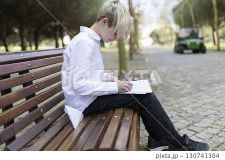 Young student writing on paper while sitting on bench in park Young student writing on paper while sitting on bench in park 130741304