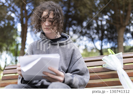 Young man reading documents on park bench with folded umbrella Young man reading documents on park bench with folded umbrella 130741312